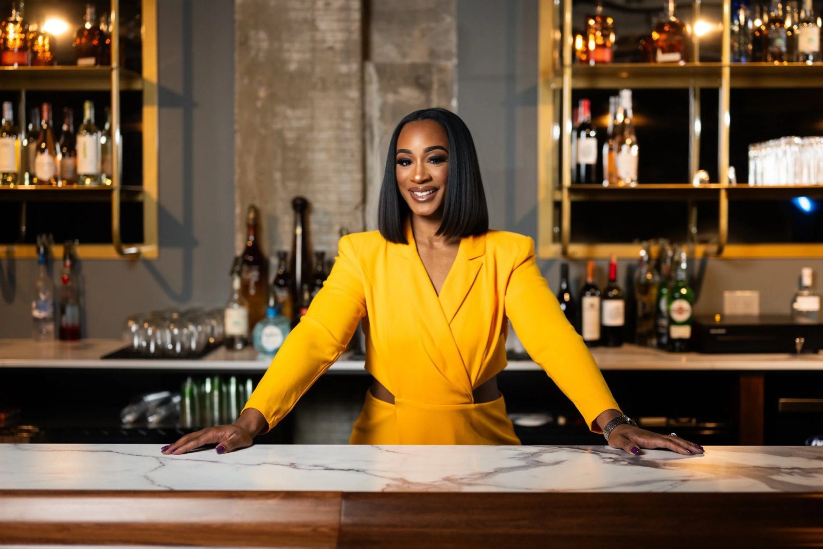 Lauren Daniels of Sumptuous Spirits in a yellow suit behind a marble bar