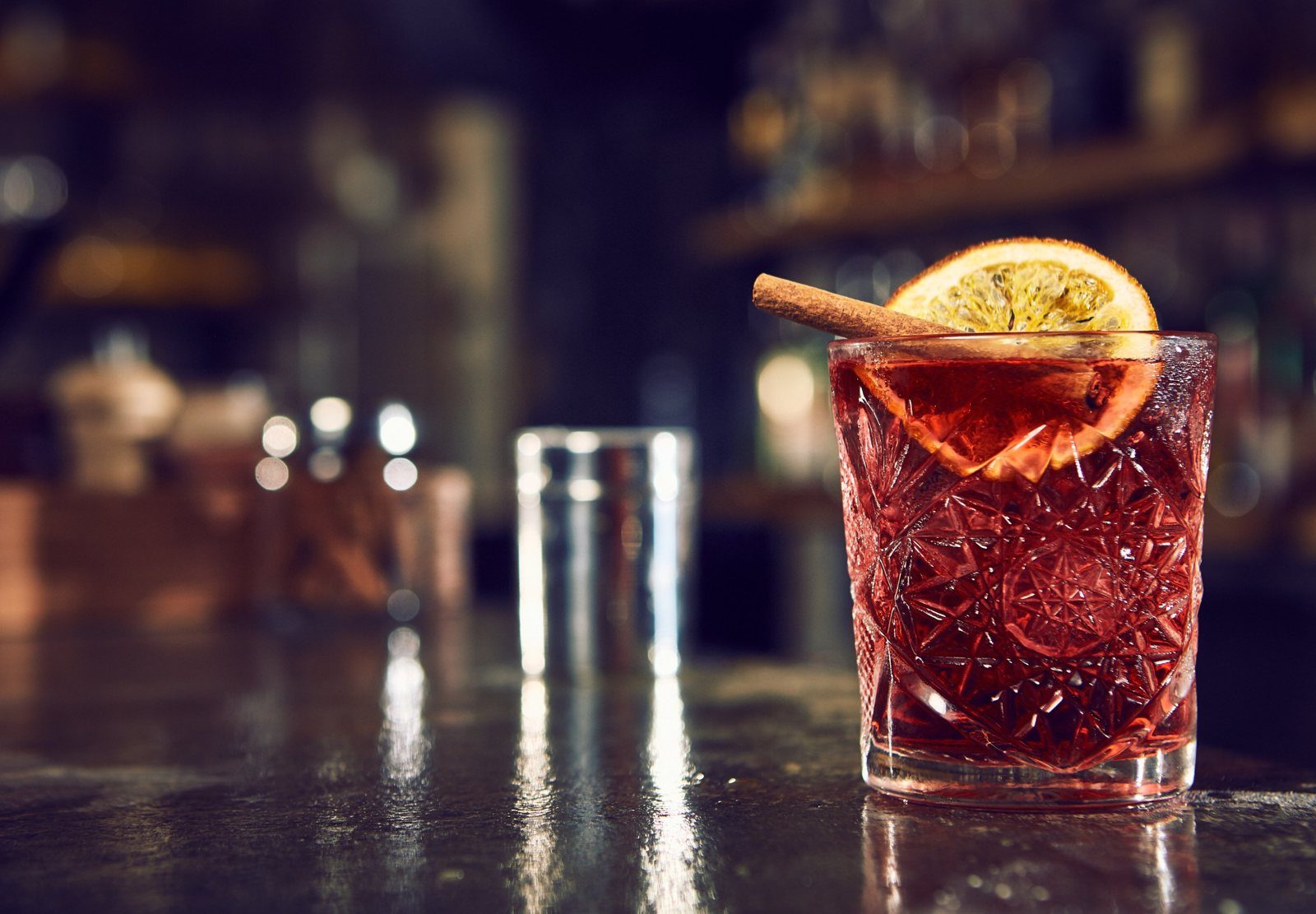 A red cocktail with an orange slice and cinnamon stick on a bar